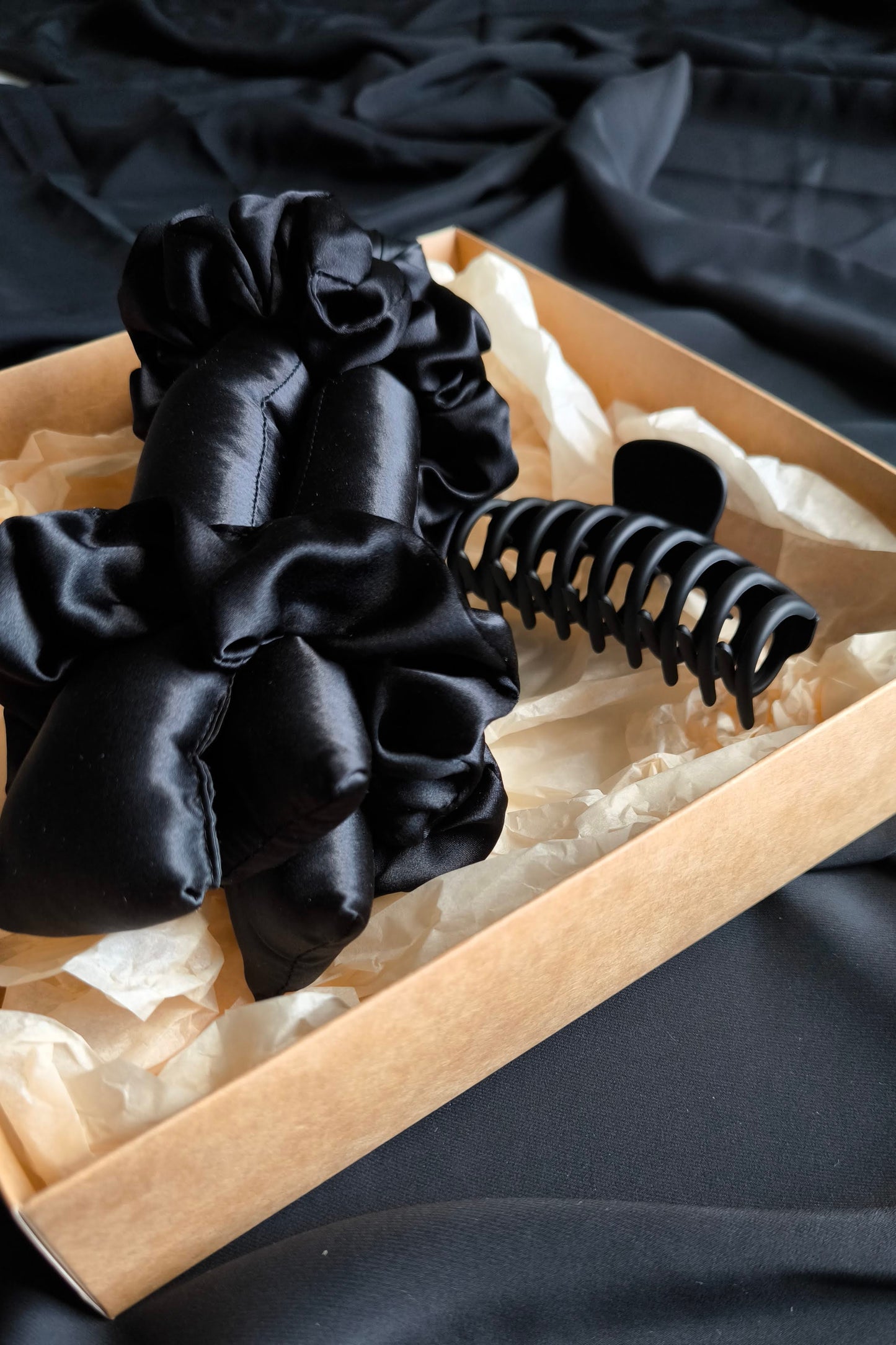 Black satin hair scrunchies and a black heatless curler in a box on a dark fabric background