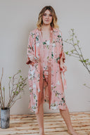 A woman standing in an interior setting wearing a kimono-style long robe with a floral pattern in peach tones.