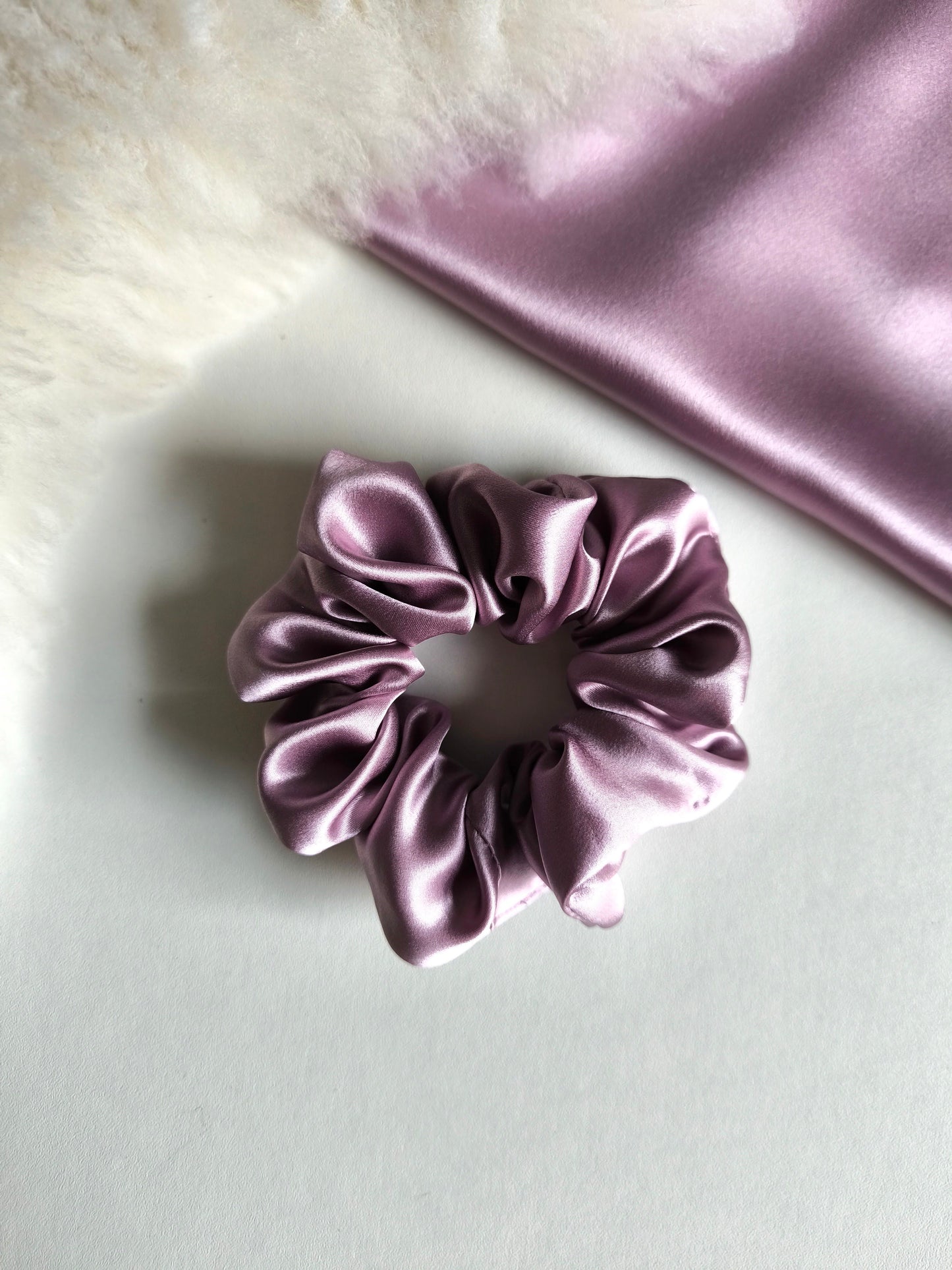 Purple satin scrunchie on a light gray surface with a piece of purple satin fabric in the background.