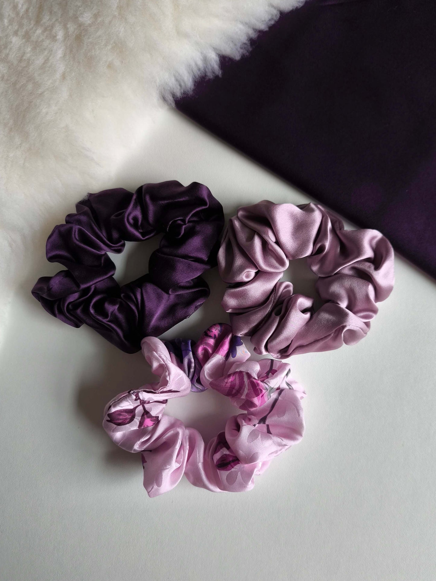 Three purple and pink scrunchies on a white surface with a textured background.