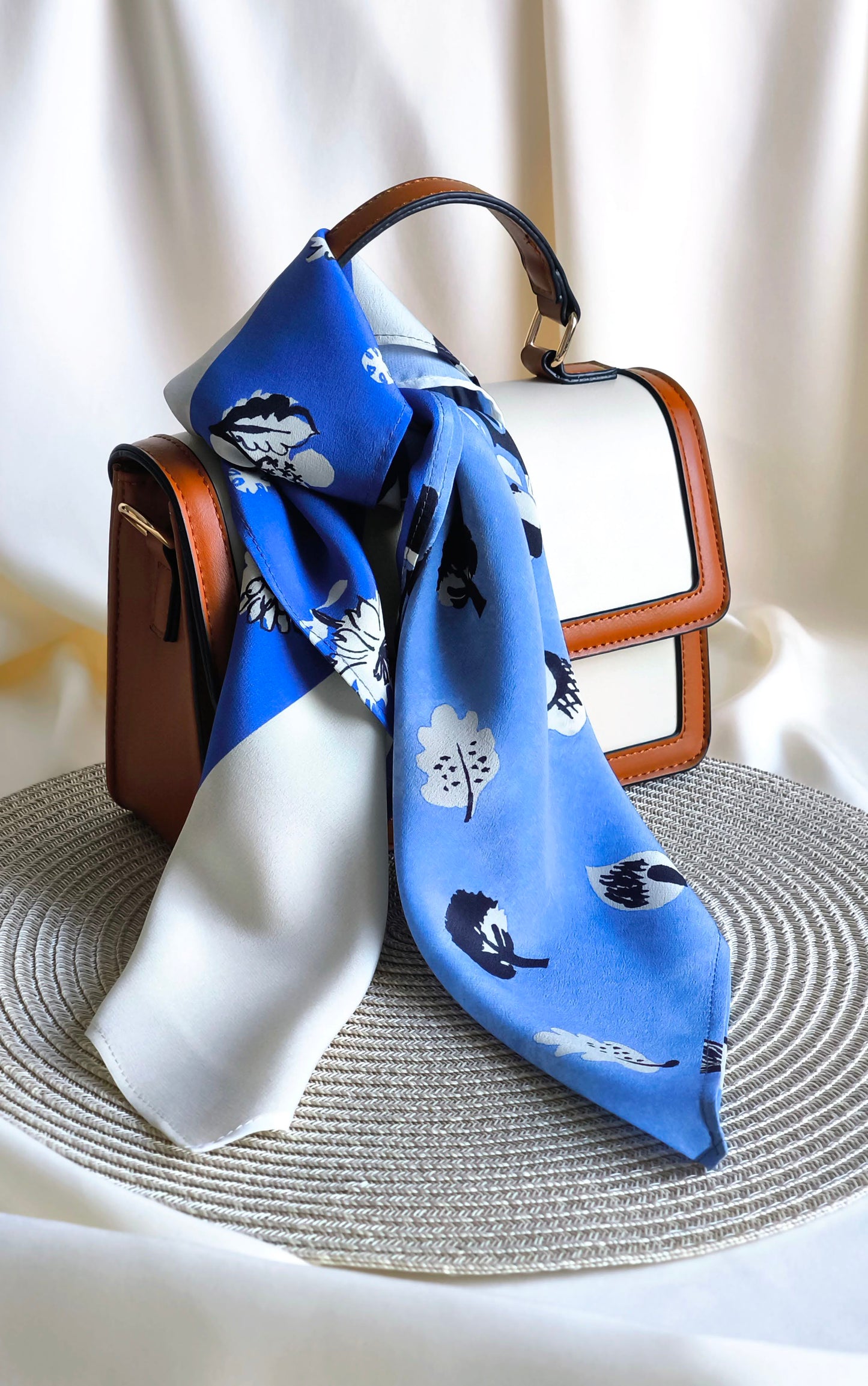 Blue floral scarf draped over a brown handbag on a textured surface with a white background