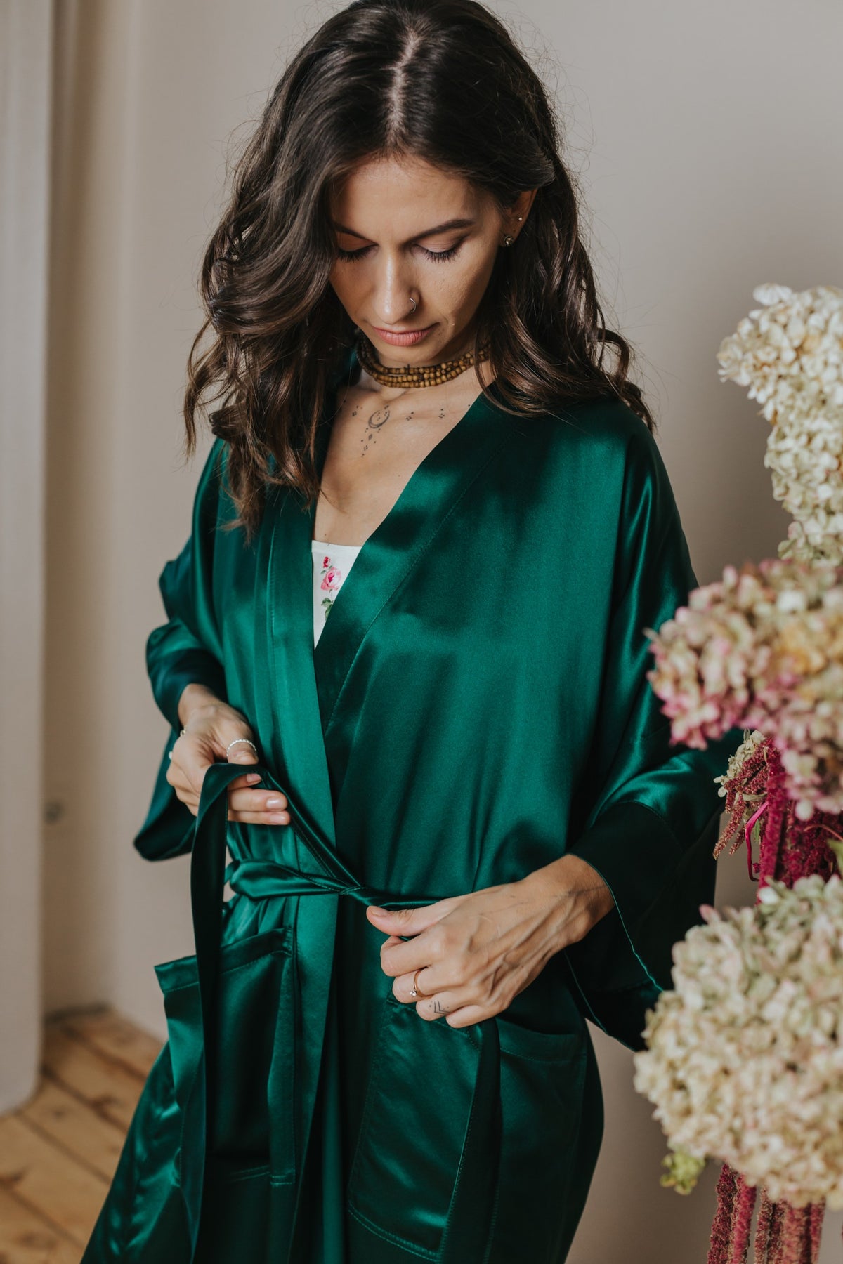 A woman wearing a shiny emerald green silk robe, tying the waist belt, with dried flowers on the right side of the frame.