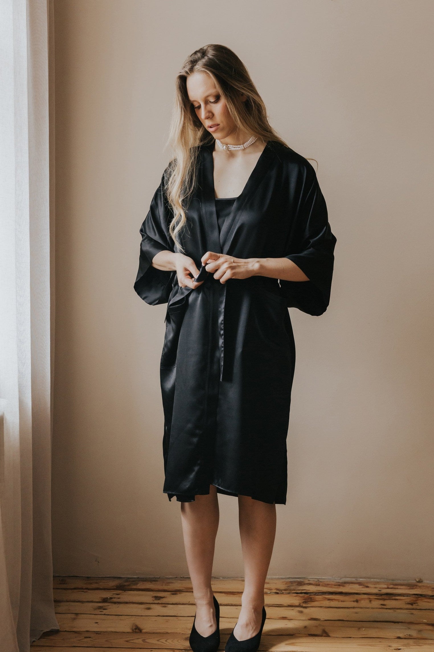 A woman wearing a black silk robe with wide sleeves and a button front, standing in a room with a beige wall and wooden floor.
