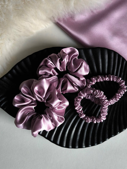 Set of pink satin hair scrunchies on a black textured surface with a soft pink fabric background.