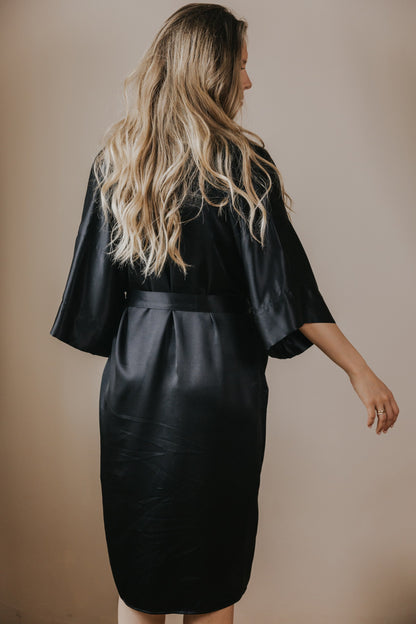A woman with long blonde wavy hair is shown from behind wearing a black silk robe with wide three-quarter sleeves and a matching waist tie, set against a plain beige studio background.
