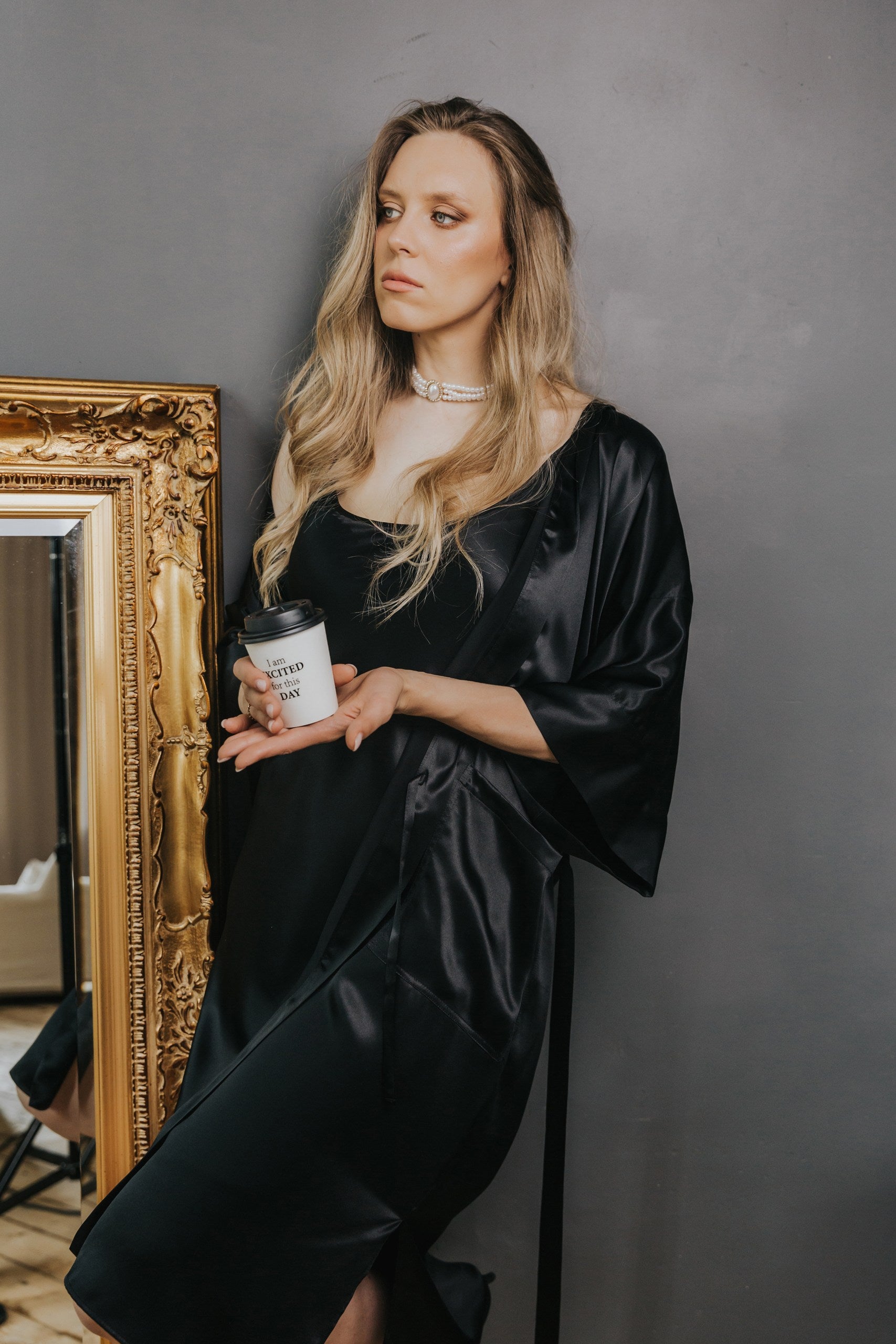 A woman with blonde hair wearing a black satin robe and holding a coffee cup, standing next to a gold-framed mirror against a gray wall.