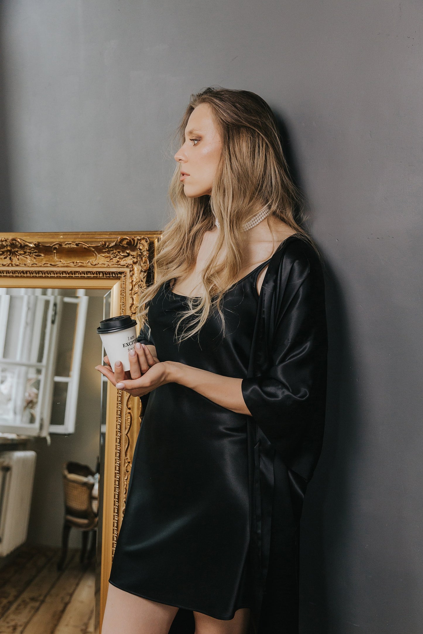 A woman with blonde hair stands against a dark gray wall, wearing a black satin slip dress and a matching black satin robe while holding a white coffee cup. An ornate gold mirror is visible on the left.