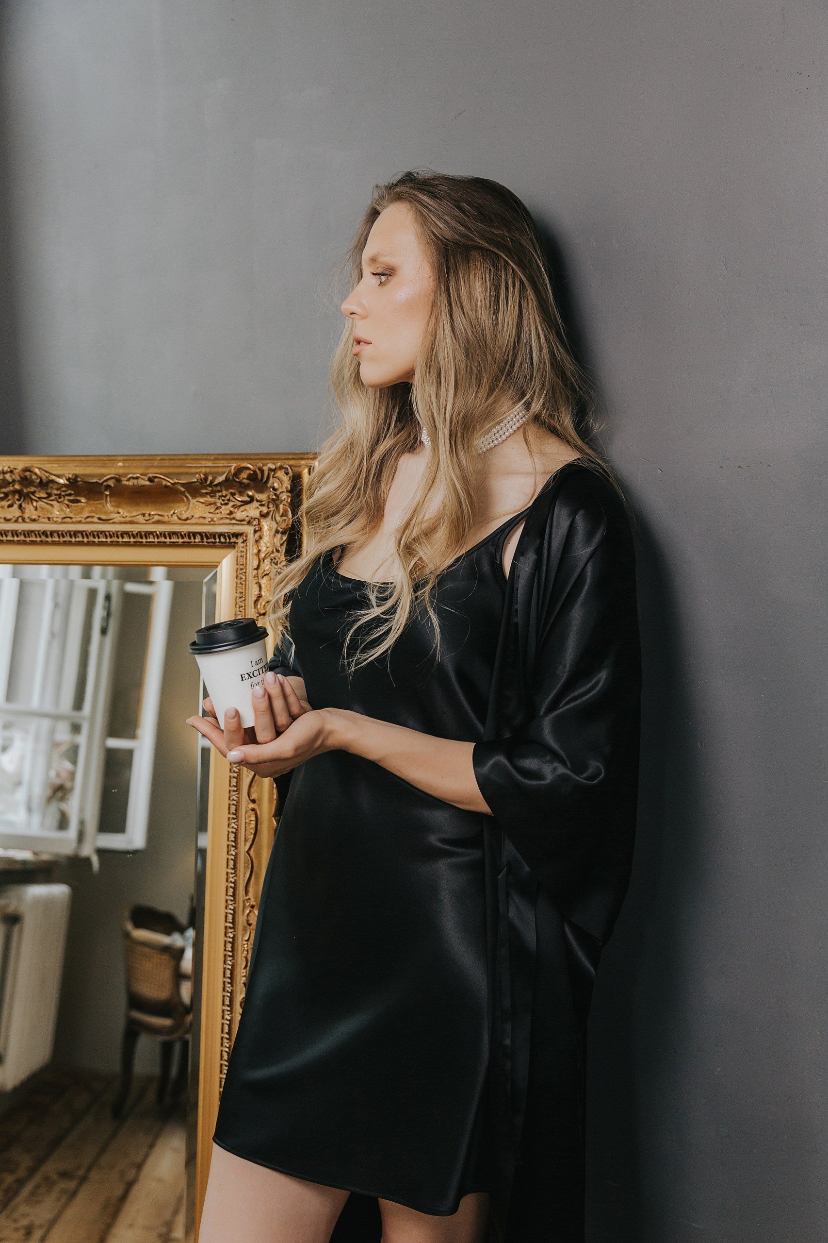 A woman with blonde hair stands against a dark gray wall, wearing a black satin slip dress and a matching black satin robe while holding a white coffee cup. An ornate gold mirror is visible on the left.