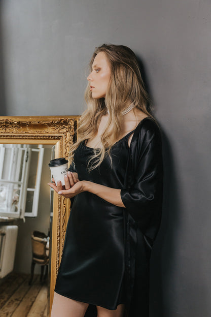 A woman with blonde hair stands against a dark gray wall, wearing a black satin slip dress and a matching black satin robe while holding a white coffee cup. An ornate gold mirror is visible on the left.