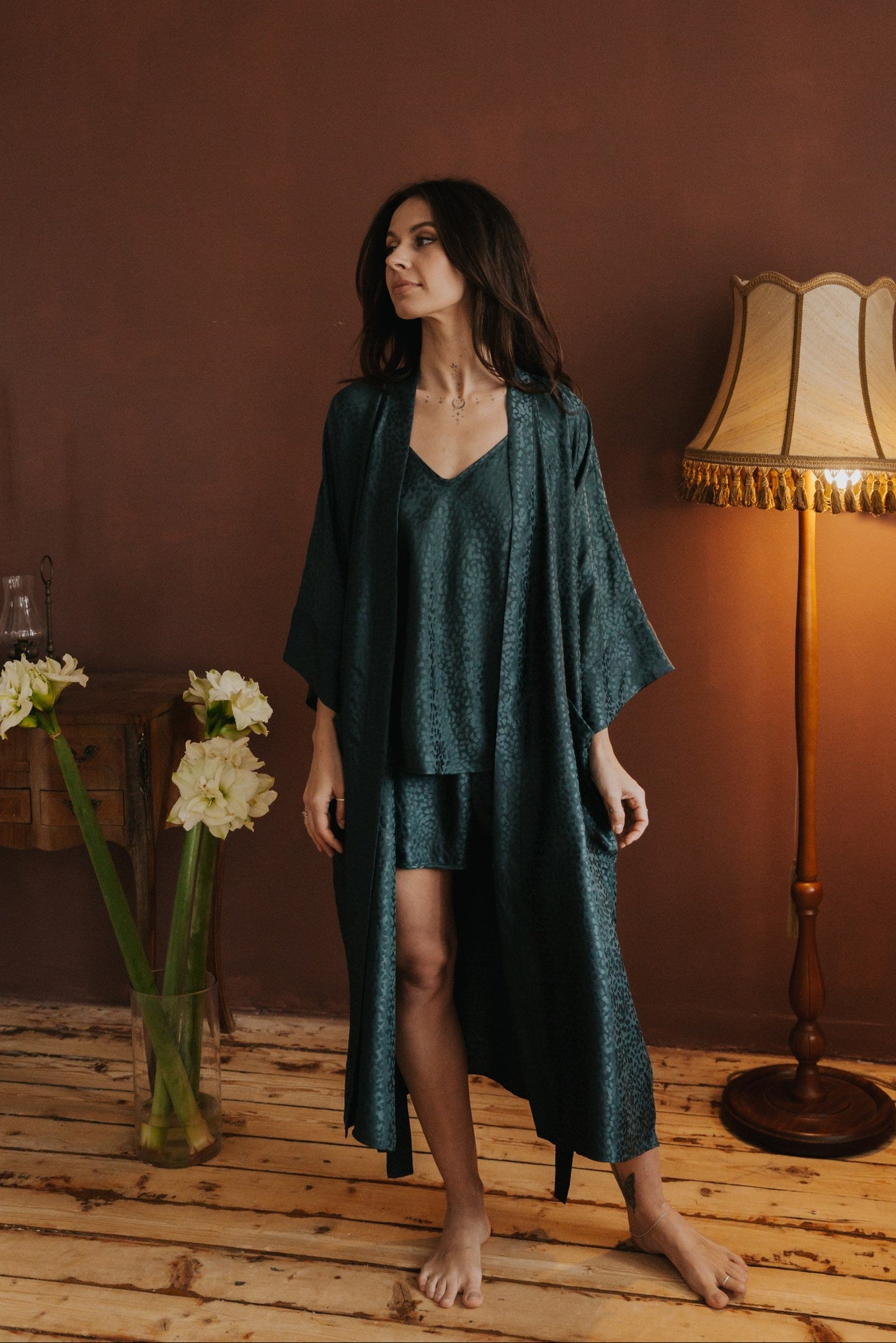 Woman in a dark green outfit standing in a room with a lamp and flowers.