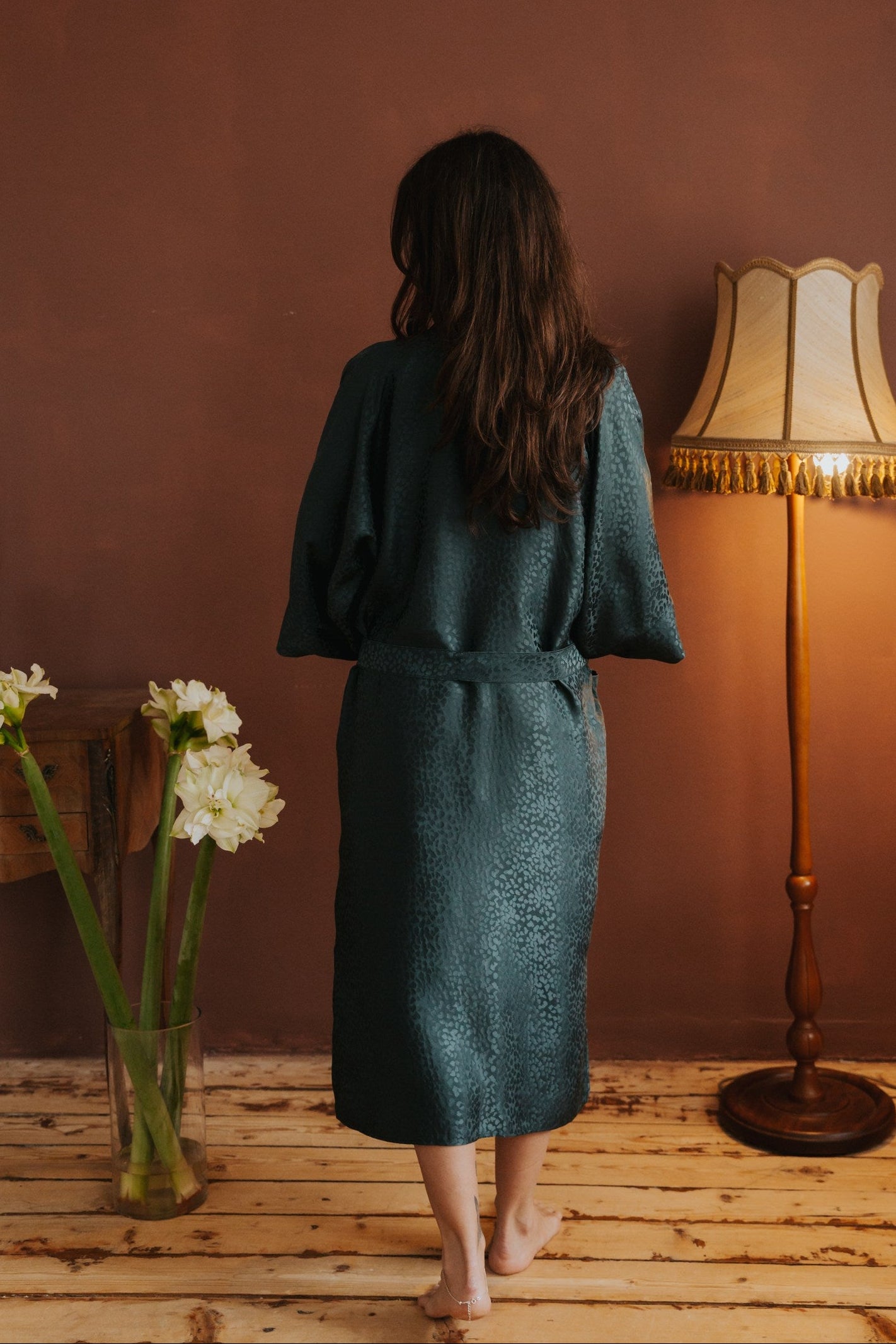 Woman in a green robe standing in a room with a lamp and flowers on a wooden floor.