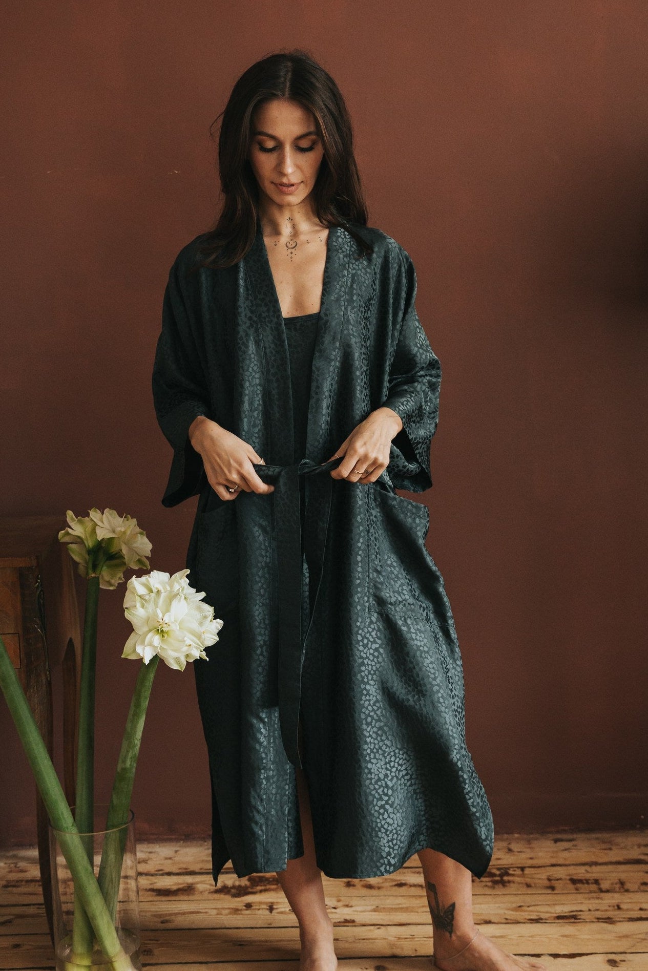 Woman in a dark green robe standing in a room with a vase of flowers on a wooden floor.
