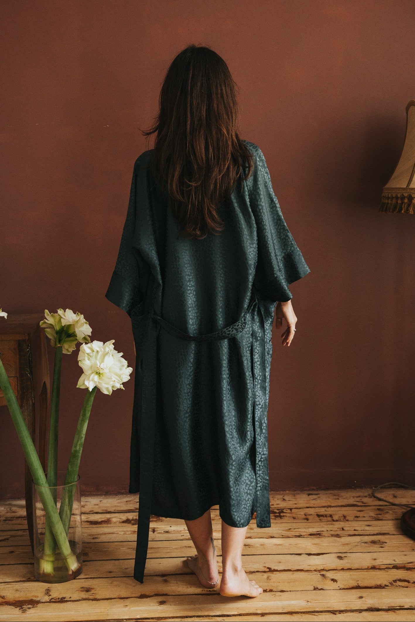 Woman in a dark green dress standing in a room with a vase of flowers and a lamp.