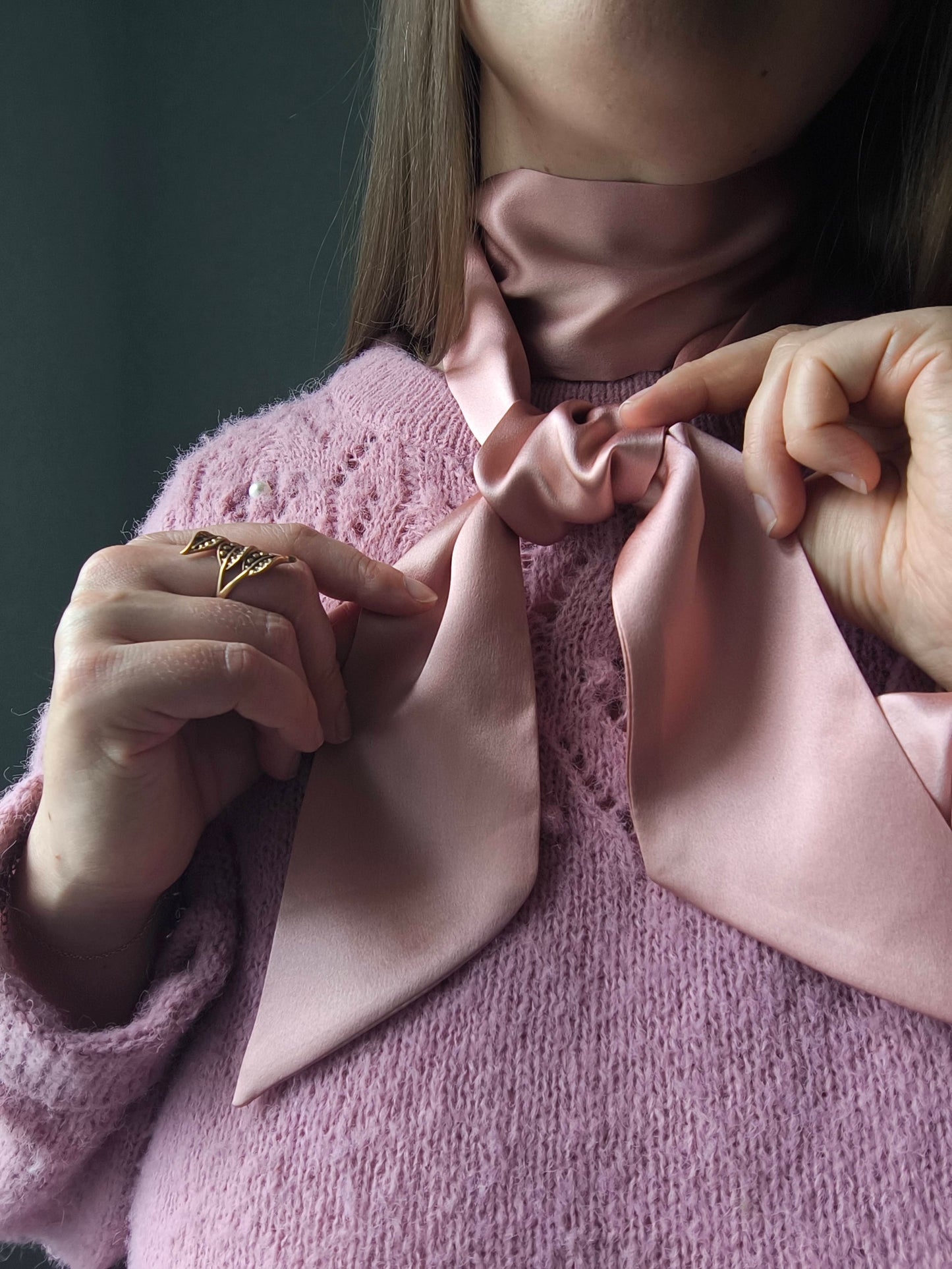 Person adjusting a pink silk scarf on a matching pink sweater against a dark background