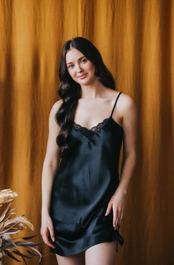 A woman standing in a black silk nightgown with lace details, against an orange background.