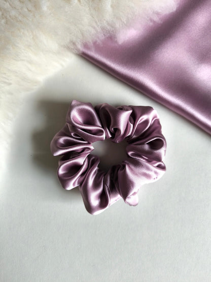 Purple satin scrunchie on a light gray surface with a piece of purple satin fabric in the background.