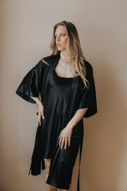 A woman modeling a black silk robe over a matching black satin slip dress, standing against a solid beige studio background.