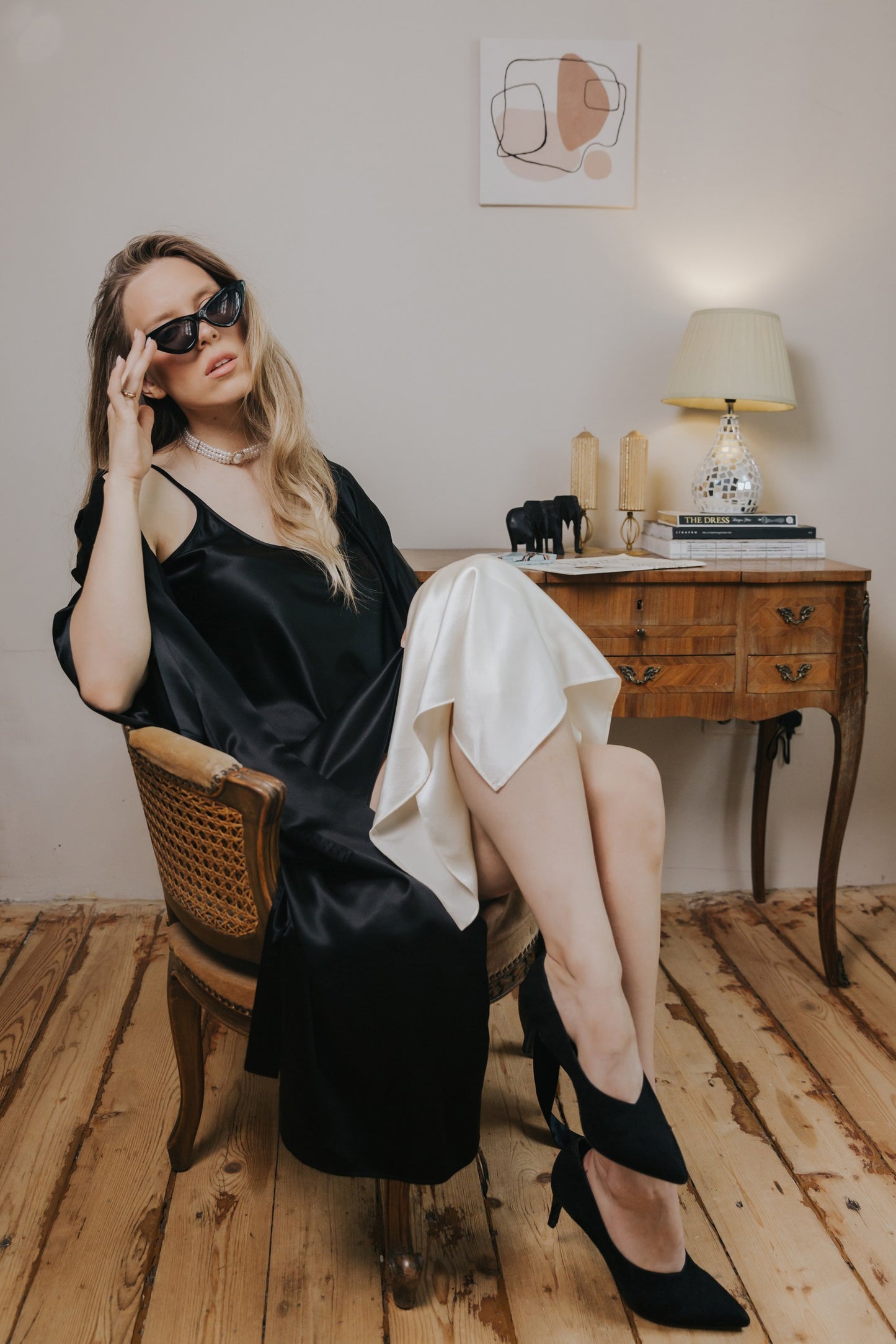 A woman wearing a black satin slip dress and black sunglasses sits in a wooden chair in a vintage room with a wooden desk, lamp, and wood floor.