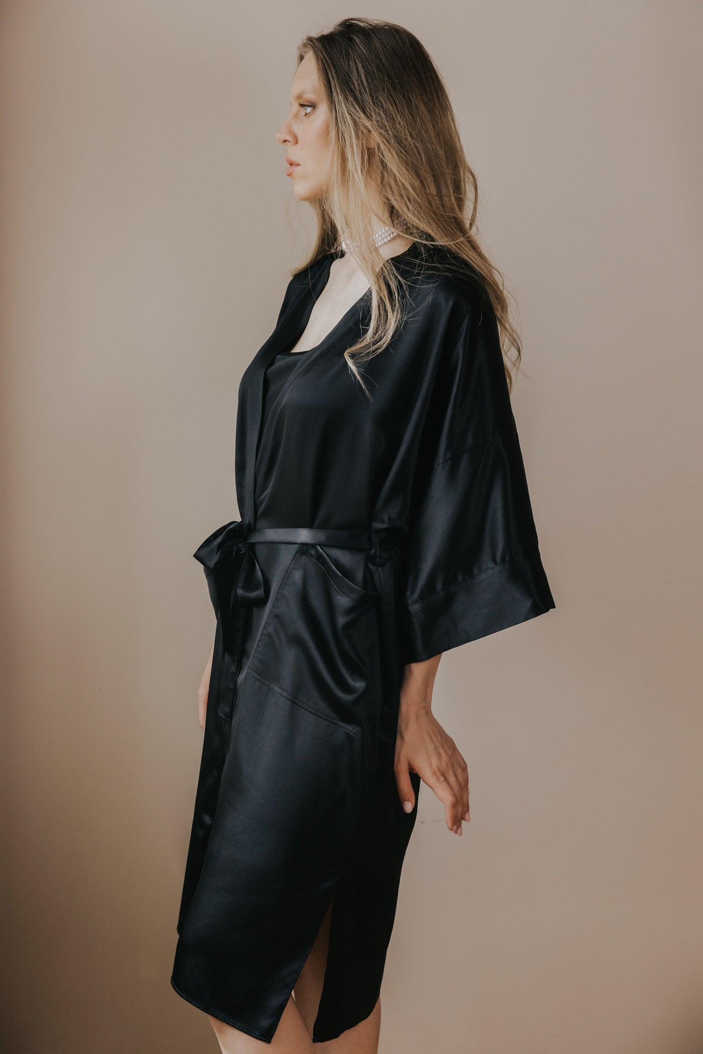 A woman wearing a black silk satin robe with wide sleeves and a self-tie belt, shown in profile against a plain beige studio background.