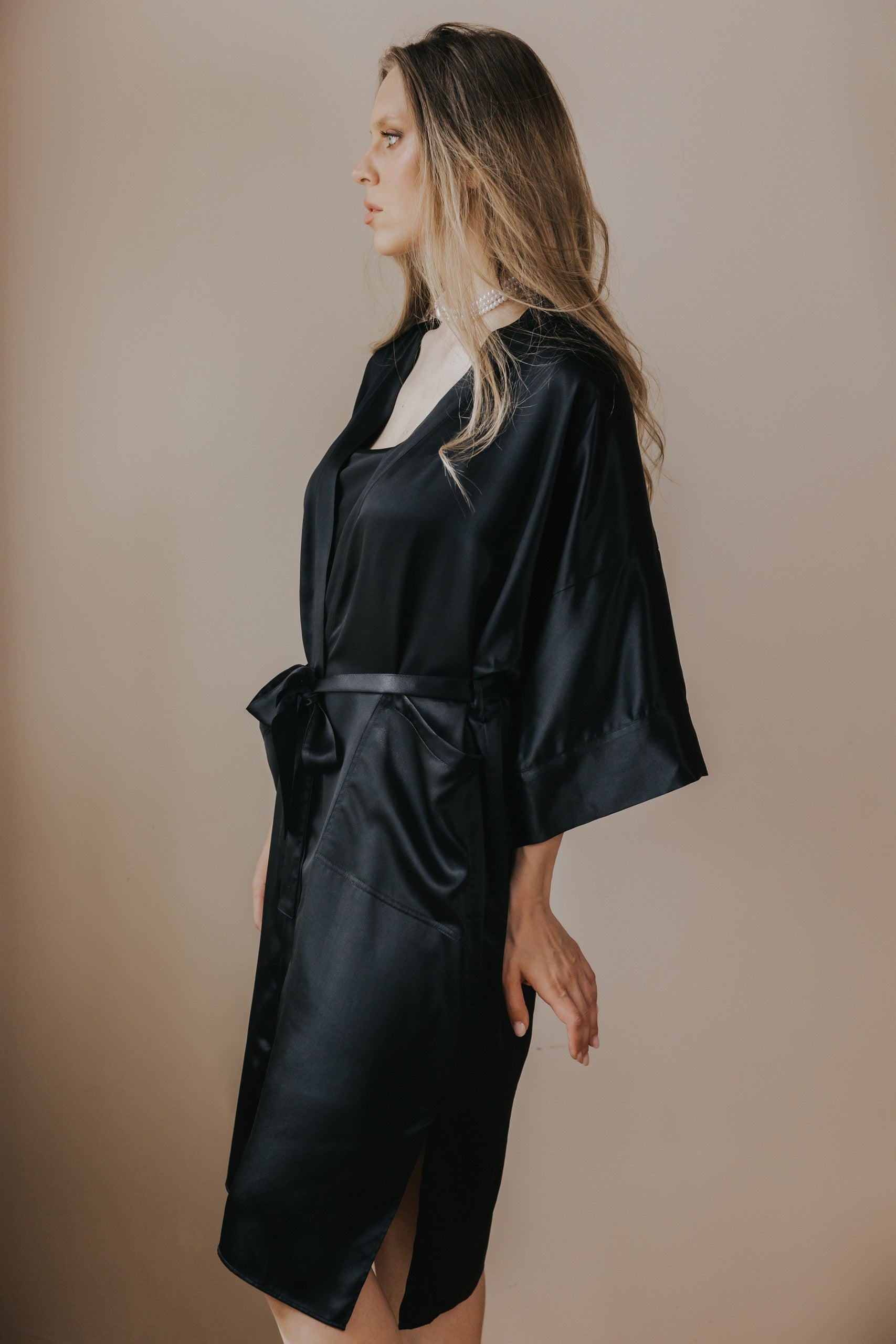 A woman wearing a black silk satin robe with wide sleeves and a self-tie belt, shown in profile against a plain beige studio background.