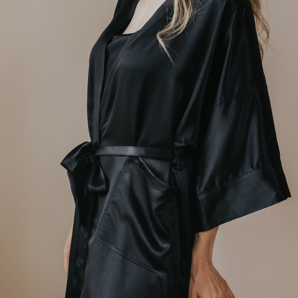 A woman wearing a black satin robe with wide sleeves and a self-tie belt, shown in profile against a plain beige studio background.